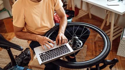 Young casually clothed man using laptop while repairing his bike at home