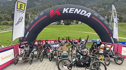 Gruppe von Menschen in geländegängigen Handbikes unter einem KENDA-Startbogen bei einem Outdoor-Event in den Bergen.