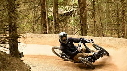 Eine Person mit Helm fährt ein geländegängiges Liegedreirad (Handbike) durch eine Kurve auf einem Schotterweg im Wald.