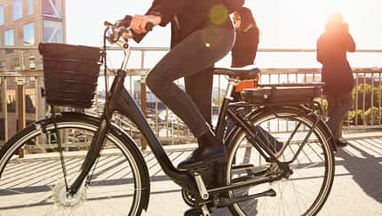 Low section of mature woman riding electric bicycle by commuters on bridge in city against sky