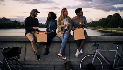 Vier Freunde essen Pizza auf einer Brücke bei Sonnenuntergang, mit Fahrrädern im Vordergrund.
