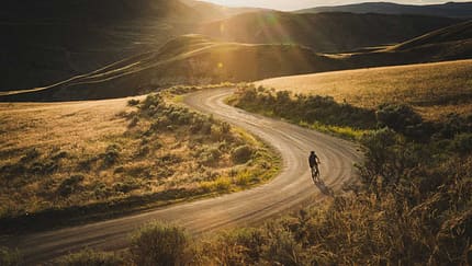 Gravelbiker fährt bei Sonnenuntergang eine staubige Serpentinenstraße durch hügelige Landschaft


