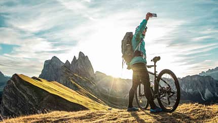 Mountainbikerin macht ein Selfie am Gipfel mit atemberaubender Bergkulisse bei Sonnenaufgang.


