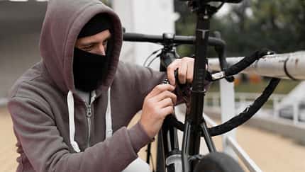 Caucasian male Thief in balaclava and hood breaks the lock on a bicycle in the street during the day. Closed face and hacking. Stealing bicycles.