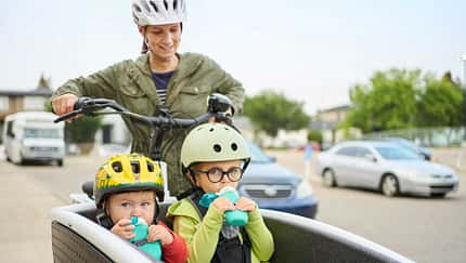 Mutter fährt zwei Kinder im Lastenrad durch ein Wohngebiet, beide Kinder mit Helmen und Trinkflaschen
