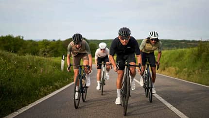 Drei Rennradfahrer fahren in Teamformation auf einer offenen Landstraße, flankiert von grüner Natur.