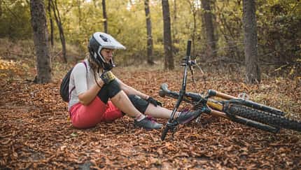 Eine Mountainbikerin sitzt in einem Wald mit herabgefallenem Herbstlaub, nachdem sie vom Fahrrad gefallen ist und sich verletzt hat. Eine Mountainbikerin sitzt in einem Wald mit herabgefallenem Herbstlaub, nachdem sie vom Fahrrad gefallen ist und sich verletzt hat.