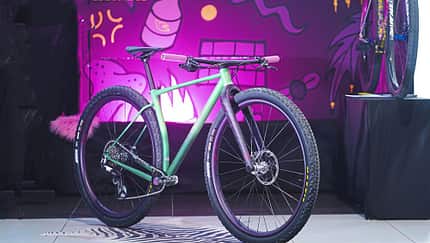 Ein modernes Mountainbike mit farbigem Rahmen und breiten Stollenreifen steht auf einer Ausstellungsfläche vor einer bunt beleuchteten Wand mit Comic-Grafik. Das Fahrrad ist komplett aufgebaut und wird prominent präsentiert.