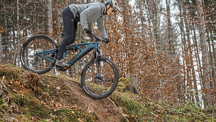 Ein Mountainbiker fährt mit einem E-Mountainbike einen steilen Abhang im Wald hinab. Das Fahrrad ist ein vollgefedertes E-MTB, der Fahrer trägt Helm und Sportbekleidung.