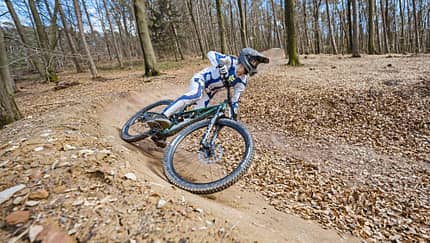 Mountainbiker in voller Schutzausrüstung fährt auf einem Trail im Wald durch eine Steilkurve.