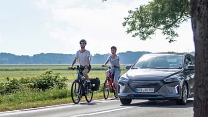 Zwei Radfahrer fahren auf einer Landstraße und werden von einem Auto überholt 