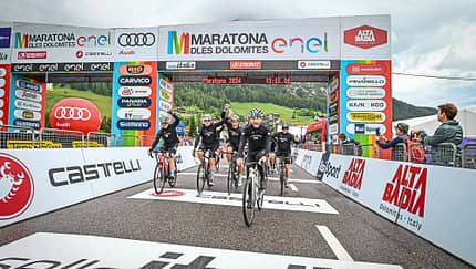Gruppe von Radfahrern überquert die Ziellinie beim Maratona dles Dolomites 2024 unter dem offiziellen Zielbogen mit Sponsorenlogos.