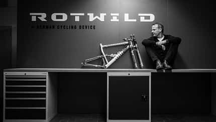 Schwarzweiß-Foto einer Person, die auf einer Werkbank neben einem Fahrradrahmen sitzt. Im Hintergrund das Logo 'Rotwild' und der Schriftzug 'German Cycling Device'.