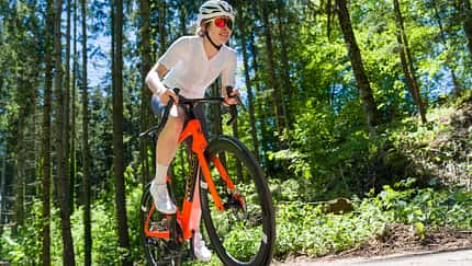 Frontalansicht einer Radsportlerin auf dem orangenen Storck Fascenario.5 in Fahrt auf einer Landstraße im Wald. Frontalansicht einer Radsportlerin auf dem orangenen Storck Fascenario.5 in Fahrt auf einer Landstraße im Wald.