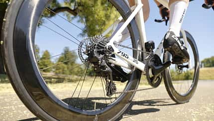 Look 795 Blade RS, Look-Rennrad, Aero-Rennrad, Carbonrahmen, Scheibenbremse, Kettenschaltung, Schaltwerk, Aero-Laufrad, Rennrad-Antrieb, Straßenradsport Die Aufnahme zeigt das Rennrad Look 795 Blade RS aus der Perspektive des Hinterrades während der Fahrt. Sichtbar sind der Carbonrahmen, eine Kassette mit Schaltwerk, tiefprofilige Aero-Laufräder sowie eine Scheibenbremse am Hinterrad. Der Fokus liegt auf dem Antrieb und der Bremsanlage auf einer sonnigen Straße im Freien.