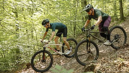 Das Foto zeigt zwei Mountainbiker auf Hardtail-Bikes bei der Abfahrt über einen schmalen, laubbedeckten Singletrail im Wald. Die Räder besitzen Federgabeln, große 29‑Zoll-Reifen und Scheibenbremsen; die Fahrer tragen Helme, Handschuhe und teils Knieprotektoren. Das Datum 10.08.2026 lässt sich aus dem Dateinamen ableiten.