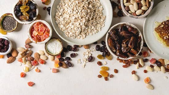 Variety of dried fruits. nuts. honey and oat flakes in ceramic bowls for cooking homemade healthy breakfast muesli or granola energy bars over white texture background. Flat lay. space.