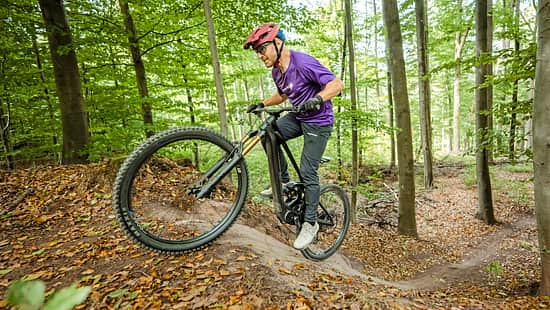 Das Foto zeigt einen Fahrer auf einem Mountainbike auf einem Wald‑Singletrail bei Schorndorf, aufgenommen am 15.09.2026 laut Dateiname. Das Rad ist als E‑Mountainbike mit Mittelmotor und integriertem Akku im Unterrohr erkennbar; eine lange Federgabel und grobstollige Reifen unterstützen die Bergauffahrt.