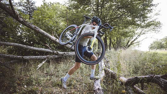 Das Foto zum Thema „Cyclo vs Gravel“ zeigt eine Person, die ein Dropbar-Geländerad (Cyclocross/Gravel-Bike) über umgestürzte Baumstämme in einem Wald trägt. Sichtbar sind Rennlenker, Scheibenbremsen und breite Stollenreifen – eine typische Tragepassage aus dem Cyclocross, wie sie auch beim Gravel-Einsatz vorkommt.