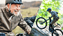 Älteres Paar mit Helmen fährt lachend auf E-Bikes durch eine hügelige Landschaft.