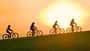 Vier Radfahrer:innen fahren bei Sonnenuntergang nebeneinander über einen Hügel – ihre Silhouetten heben sich dunkel vor dem orangefarbenen Himmel ab.