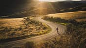 Gravelbiker fährt bei Sonnenuntergang eine staubige Serpentinenstraße durch hügelige Landschaft

