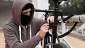 Caucasian male Thief in balaclava and hood breaks the lock on a bicycle in the street during the day. Closed face and hacking. Stealing bicycles.
