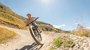Mountainbiker fährt dynamisch auf Schotterweg durch sonnige Hügellandschaft Mountainbiker fährt dynamisch auf Schotterweg durch sonnige Hügellandschaft