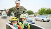 Mutter fährt zwei Kinder im Lastenrad durch ein Wohngebiet, beide Kinder mit Helmen und Trinkflaschen