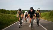 Drei Rennradfahrer fahren in Teamformation auf einer offenen Landstraße, flankiert von grüner Natur.