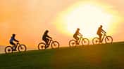Vier Radfahrer:innen fahren bei Sonnenuntergang nebeneinander über einen Hügel – ihre Silhouetten heben sich dunkel vor dem orangefarbenen Himmel ab.