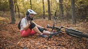 Eine Mountainbikerin sitzt in einem Wald mit herabgefallenem Herbstlaub, nachdem sie vom Fahrrad gefallen ist und sich verletzt hat.