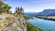 Drei Mountainbiker stehen mit ihren Fahrrädern auf einem Felsvorsprung oberhalb eines Sees mit Panoramablick auf Berge und Landschaft.