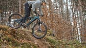 Ein Mountainbiker fährt mit einem E-Mountainbike einen steilen Abhang im Wald hinab. Das Fahrrad ist ein vollgefedertes E-MTB, der Fahrer trägt Helm und Sportbekleidung.