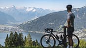 Ein Radfahrer mit Rennrad blickt auf den Zeller See und die umliegenden Berge bei Zell am See-Kaprun. Die Alpenlandschaft ist im Hintergrund deutlich sichtbar.