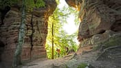 Zwei Mountainbiker fahren durch eine Felsformation im Wald bei Sonnenlicht.