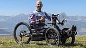 Eine Person sitzt in einem geländegängigen Handbike (Bowhead RX) auf einer Wiese vor Bergpanorama.