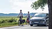 Zwei Radfahrer fahren auf einer Landstraße und werden von einem Auto überholt 