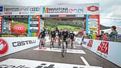 Gruppe von Radfahrern überquert die Ziellinie beim Maratona dles Dolomites 2024 unter dem offiziellen Zielbogen mit Sponsorenlogos.