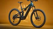 Das Bild zeigt ein Focus Jam2 Next E-Mountainbike in Studioaufnahme vor orangefarbenem Hintergrund. Das vollgefederte E-MTB ist seitlich positioniert und präsentiert Details wie den integrierten Akku, die Fox-Federgabel und grobstollige Reifen.