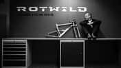 Schwarzweiß-Foto einer Person, die auf einer Werkbank neben einem Fahrradrahmen sitzt. Im Hintergrund das Logo 'Rotwild' und der Schriftzug 'German Cycling Device'.