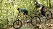 Das Foto zeigt zwei Mountainbiker auf Hardtail-Bikes bei der Abfahrt über einen schmalen, laubbedeckten Singletrail im Wald. Die Räder besitzen Federgabeln, große 29‑Zoll-Reifen und Scheibenbremsen; die Fahrer tragen Helme, Handschuhe und teils Knieprotektoren. Das Datum 10.08.2026 lässt sich aus dem Dateinamen ableiten.
