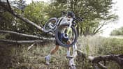 Das Foto zum Thema „Cyclo vs Gravel“ zeigt eine Person, die ein Dropbar-Geländerad (Cyclocross/Gravel-Bike) über umgestürzte Baumstämme in einem Wald trägt. Sichtbar sind Rennlenker, Scheibenbremsen und breite Stollenreifen – eine typische Tragepassage aus dem Cyclocross, wie sie auch beim Gravel-Einsatz vorkommt.
