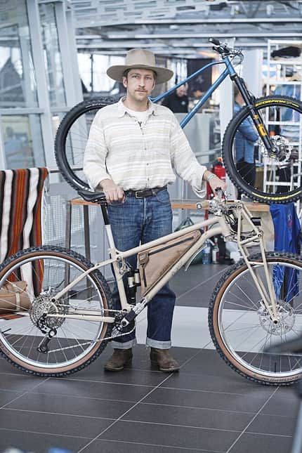 Ein Mann mit Cowboyhut steht in einer Messehalle und hält ein beiges Mountainbike mit Rahmentasche. Im Hintergrund ist ein weiteres Mountainbike ausgestellt.