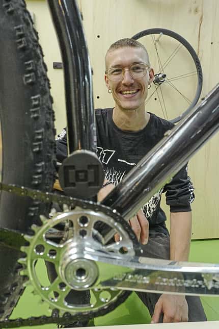 Eine Person kniet hinter einem Fahrrad und lächelt in die Kamera. Im Vordergrund sind das Tretlager, die Kurbel und der Antrieb des Fahrrads deutlich zu sehen. Im Hintergrund hängt ein Laufrad an der Wand.