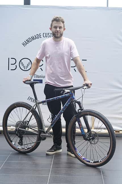 Ein Mann steht mit einem modernen Hardtail-Mountainbike vor einem Messestand mit dem Logo 'BOBUR'. Das Fahrrad hat einen Titanrahmen und eine Federgabel.