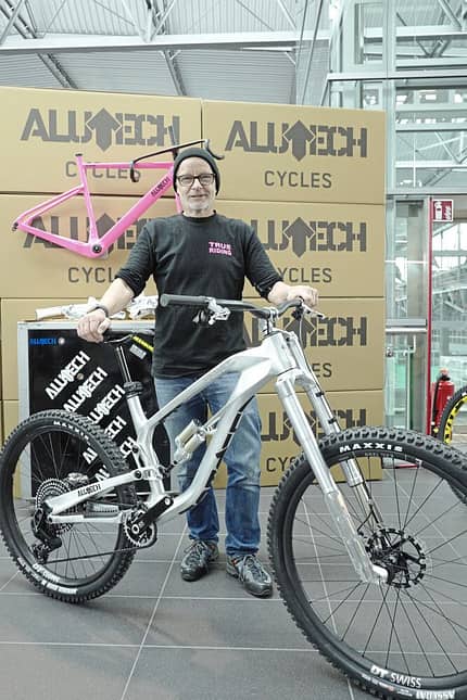Ein Mann präsentiert ein silbernes Alutech-Mountainbike vor Kartons mit Alutech-Logo. Im Hintergrund ist ein pinker Fahrradrahmen zu sehen. Die Aufnahme entstand vermutlich auf einer Messe oder in einem Showroom.
