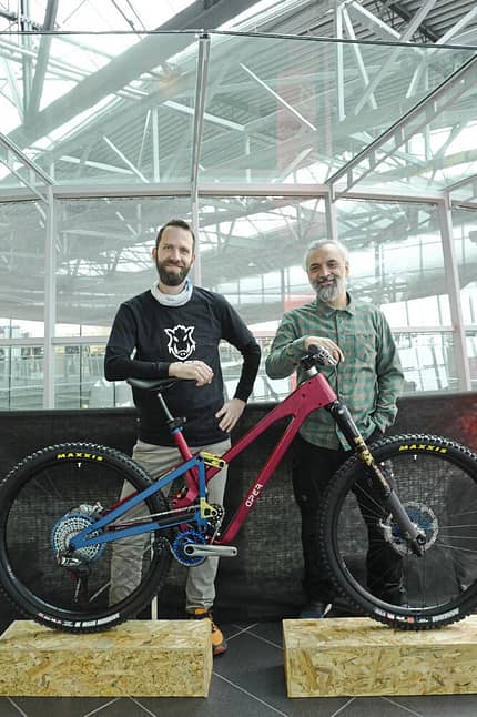 Zwei Männer stehen hinter einem modernen Mountainbike mit Vollfederung in einer Messehalle.