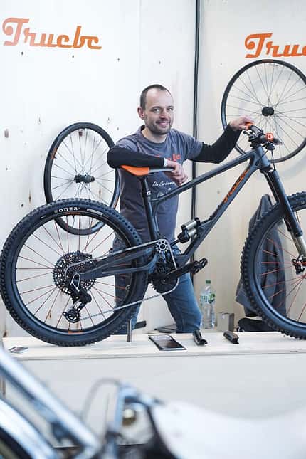 Ein Mann präsentiert ein Mountainbike am Messestand der Marke Truebc. Im Hintergrund sind zwei Laufräder an der Wand befestigt. Das Fahrrad ist mit modernen Komponenten wie Scheibenbremsen und breiten Reifen ausgestattet.