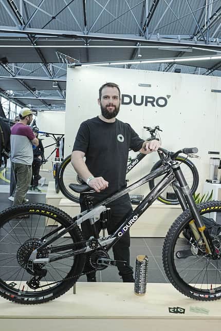 Eine Person steht auf einer Fahrradmesse neben einem modernen Mountainbike der Marke DURO. Das Fahrrad ist prominent ausgestellt, im Hintergrund sind weitere Fahrräder und Besucher zu sehen.